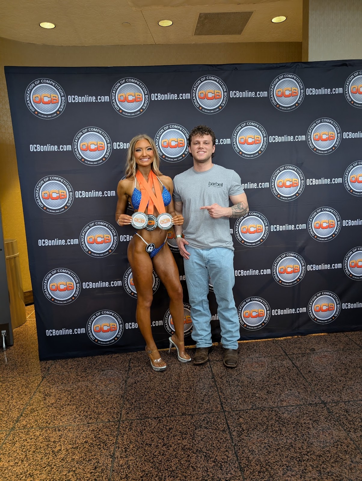 Coach Bradyn Gamber pointing proudly at his bikini athlete, who is wearing three medals at an OCB competition.